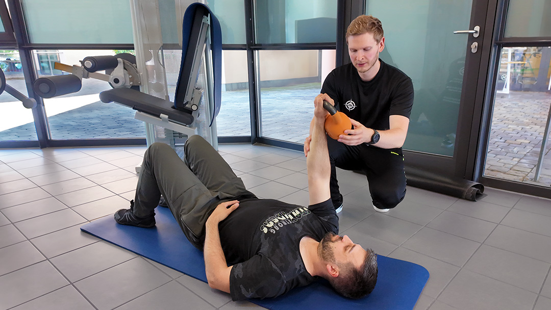 Dominik Rödl unterstützt einen auf einer Trainingsmatte liegenden Patienten bei einer Übung mit einer kleinen Hantel. Dabei führt er den ausgestreckten Arm des Patienten kontrolliert nach oben.
